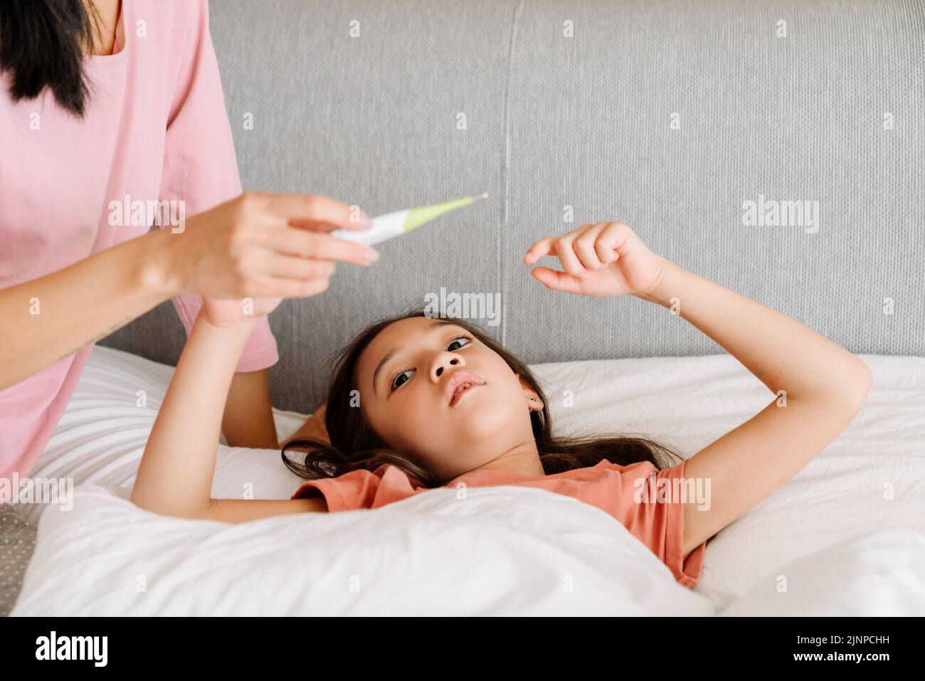 Niña asiática con gripe que mide la temperatura mientras está acostada en la cama en casa Foto de stock