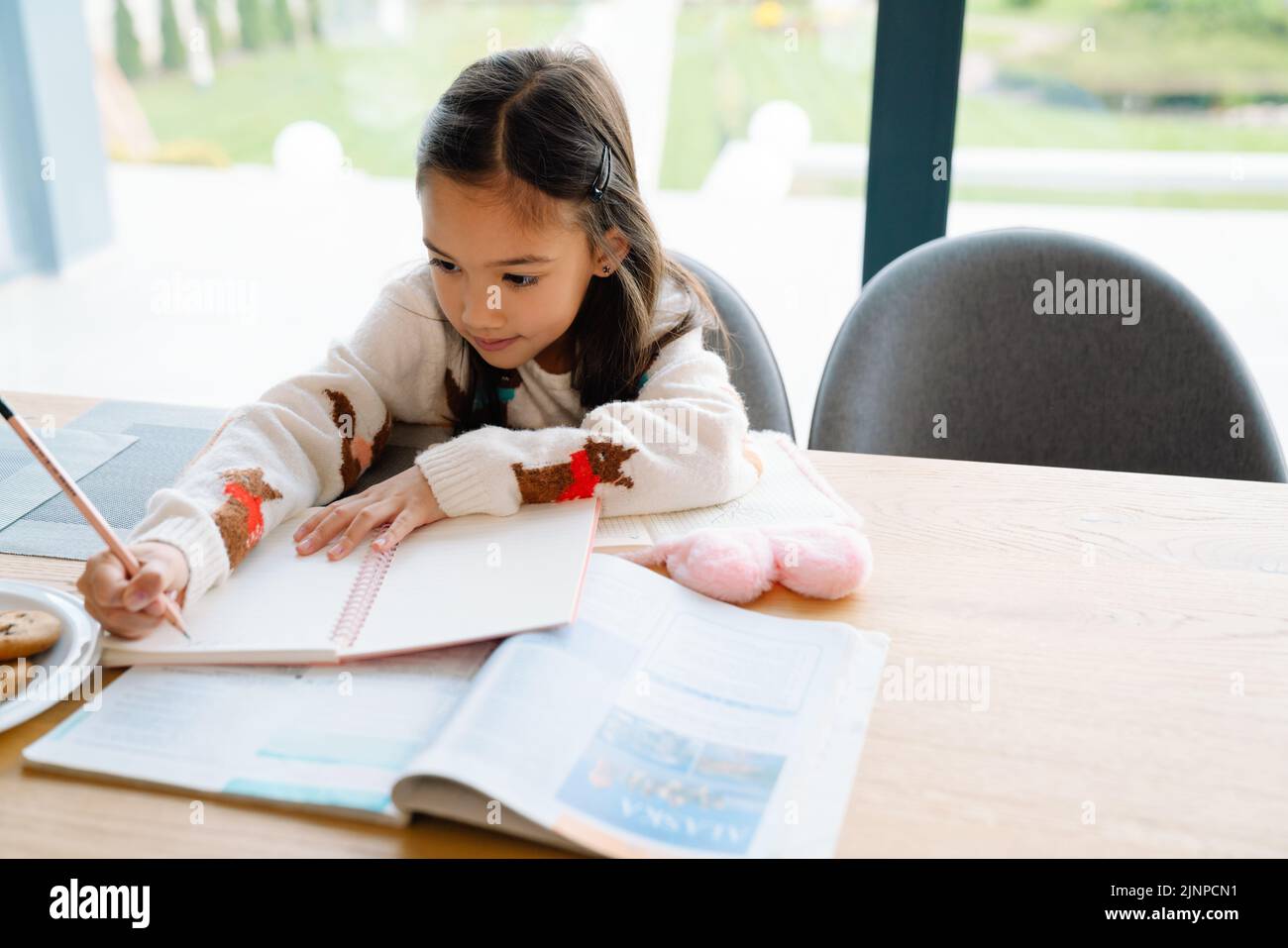 Chica asiática haciendo la tarea mientras se sienta en la mesa en casa Foto de stock
