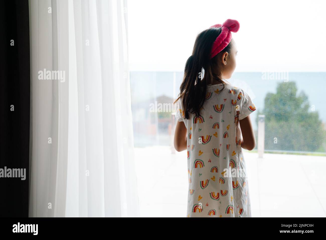 Chica asiática que usa ropa para el hogar mirando por la ventana en el dormitorio de la casa Foto de stock