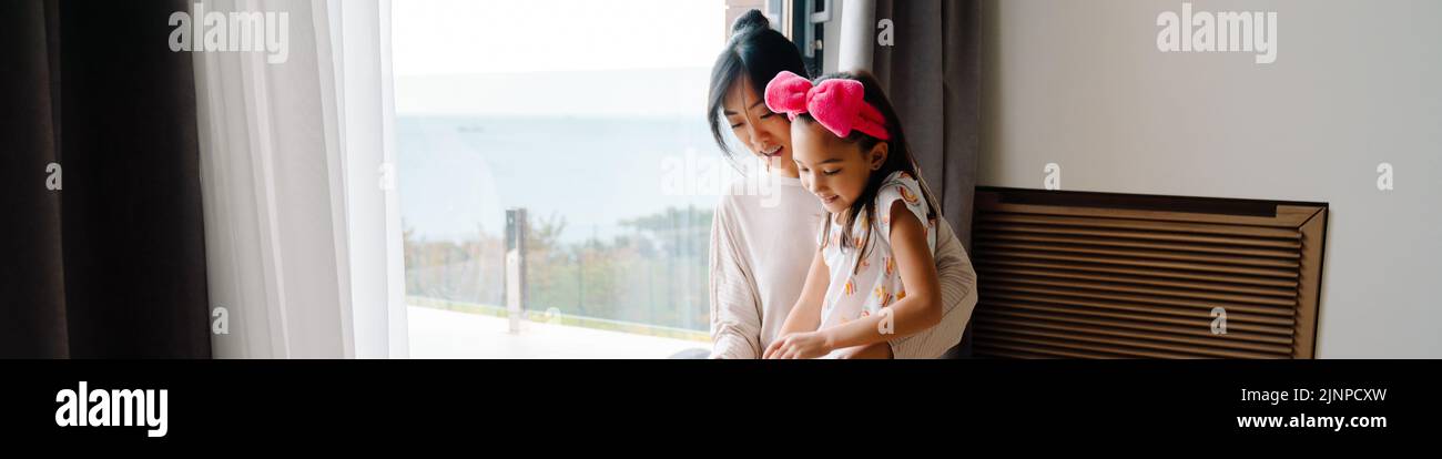 Mujer asiática y su hija hablando de pie junto a la ventana en casa Foto de stock
