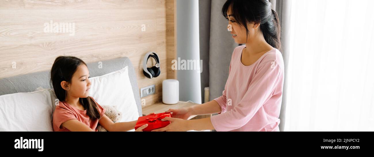Mujer asiática dando a su hija regalo mientras se sienta en la cama en casa Foto de stock