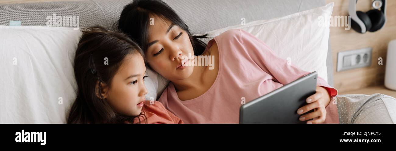 Madre asiática y su hija usando una tableta mientras se sienta en la cama en casa Foto de stock