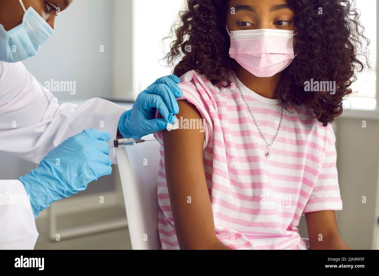 Niña afroamericana en máscara facial médica recibe la vacuna que la protegerá del virus Foto de stock