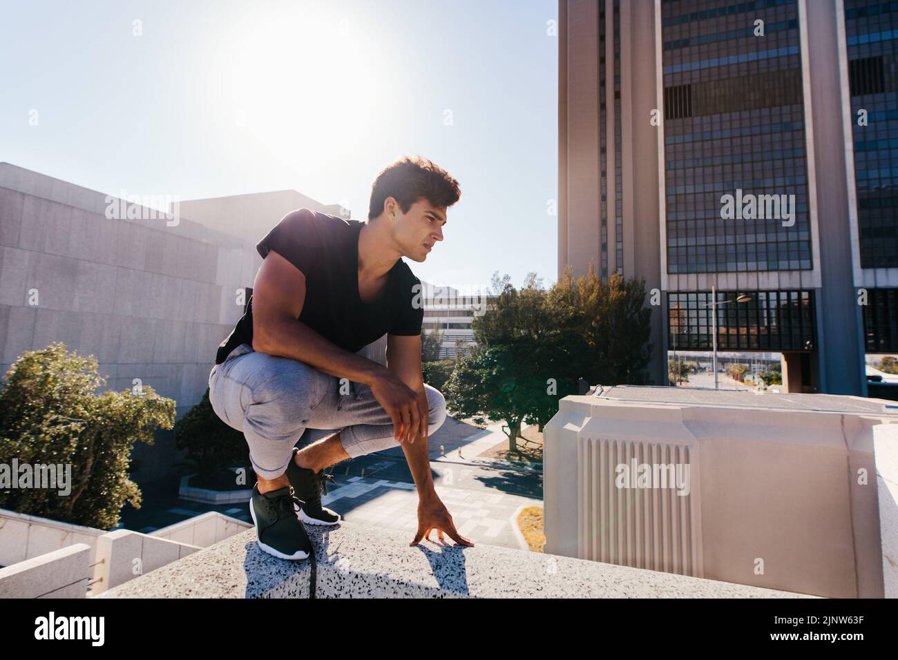 Joven deportista se pone en cuclillas al aire libre antes de demostrar las técnicas de parkour y carrera libre. El hombre joven del ajuste que se prepara para practicar freerunning en ci Foto de stock