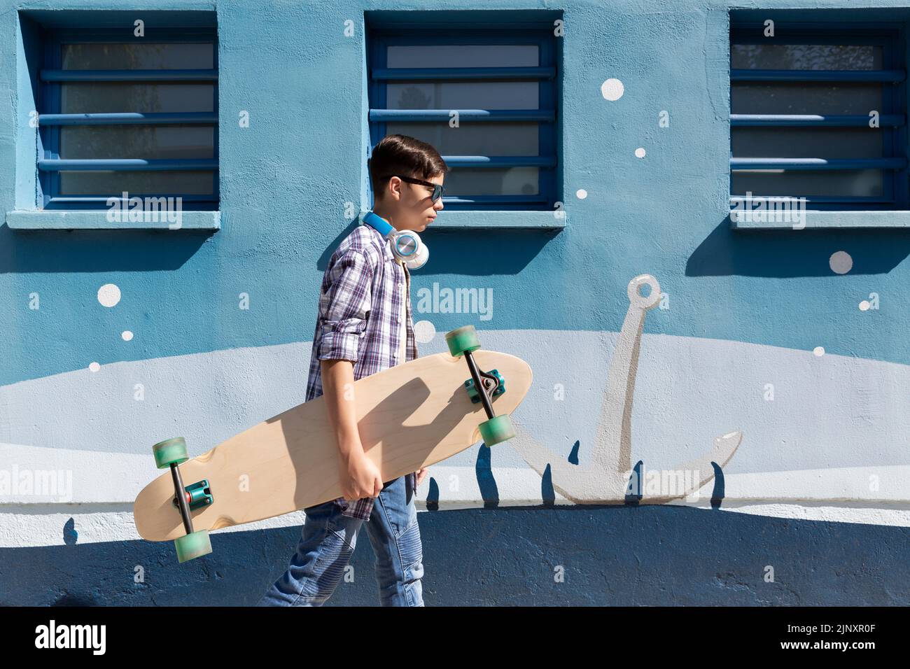 Adolescente caucásico caminando con su patineta junto a una pared azul. Foto de stock