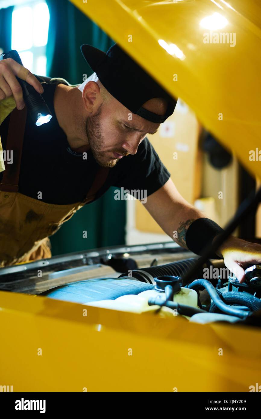 Mecánico auto serio con linterna que se agarra sobre el capó abierto del coche amarillo, comprobando defectos y piezas de fijación del motor en el garaje Foto de stock