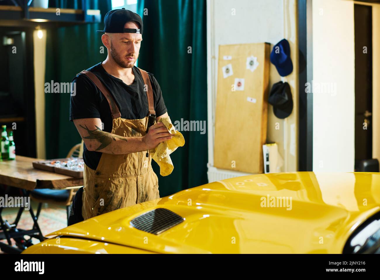 Joven reparador en ropa de trabajo frotando las manos sucias con el plumero sobre el capó de un coche eléctrico amarillo mientras se encuentra delante de él en el garaje Foto de stock