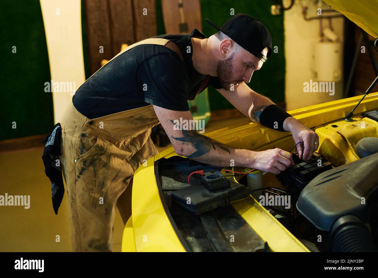 Técnico joven en ropa de trabajo que realiza diagnósticos técnicos del motor del coche mientras se dobla sobre el motor y utiliza un multímetro Foto de stock