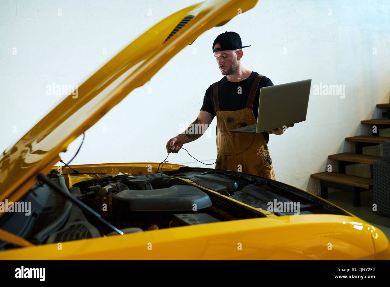 Mecánico joven con computadora portátil instalando cables en el motor del coche mientras realiza diagnósticos técnicos del motor después de la reparación Foto de stock