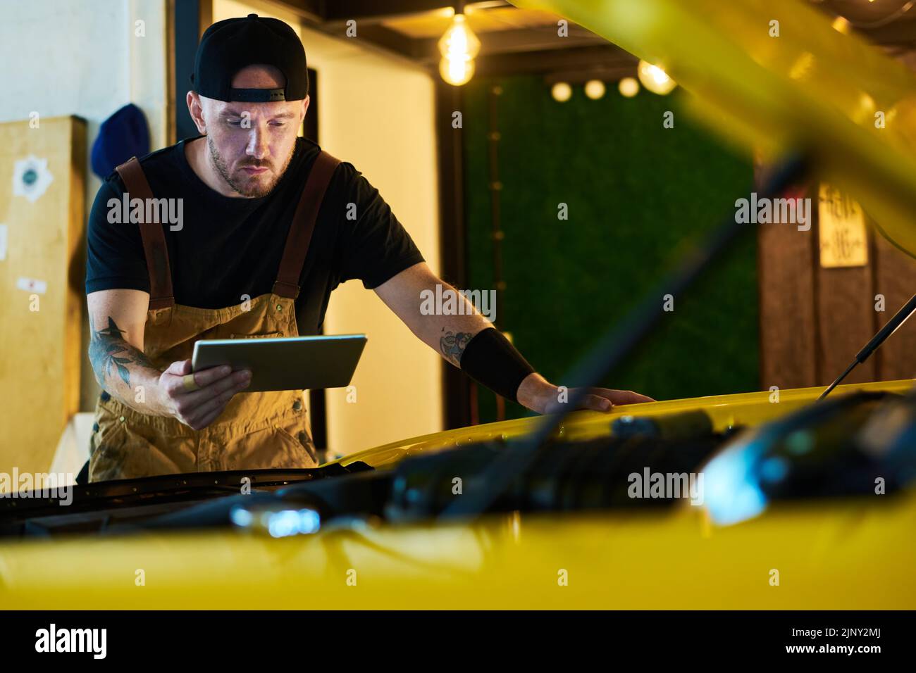 Trabajador joven del servicio de mantenimiento mirando la pantalla de la tableta mientras se dobla sobre el motor del coche y realizando una comprobación técnica Foto de stock