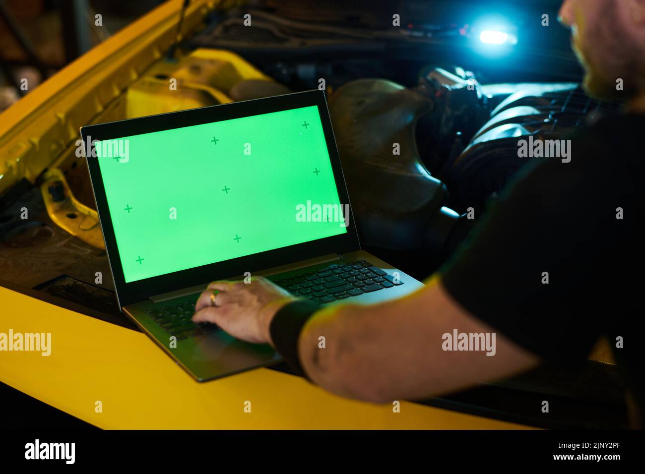 Mano de mecánico joven o reparador en el teclado del ordenador portátil con pantalla verde en el motor del vehículo con capó abierto durante el diagnóstico Foto de stock