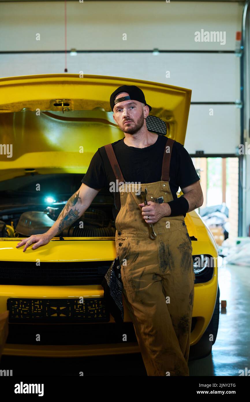 Joven trabajador serio de servicio de mantenimiento de automóviles de pie por automóvil de color amarillo con capó abierto y mirando a la cámara Foto de stock