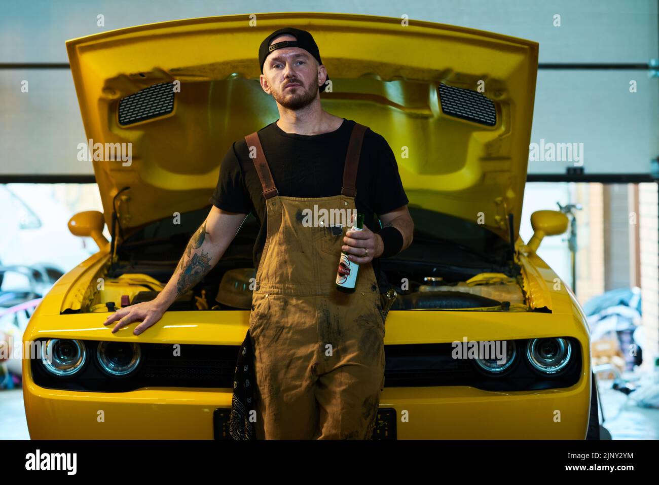 Joven técnico o ingeniero del servicio de mantenimiento con botella de cerveza parada contra coche amarillo con capó abierto en taller Foto de stock