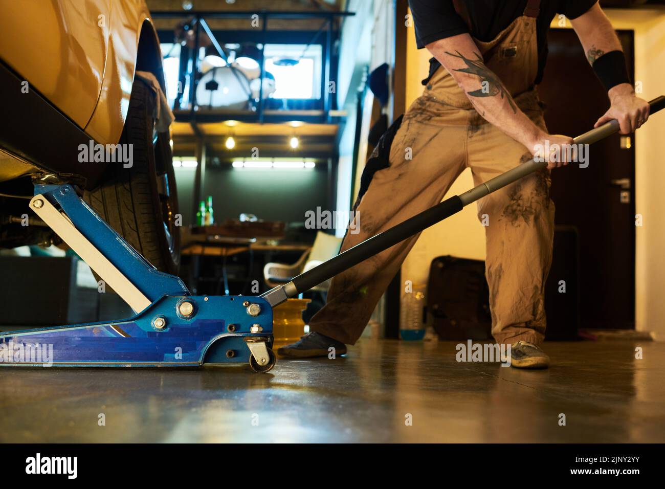 Mecánico joven en ropa de trabajo usando gato de tijera para levantar el coche sobre el piso antes de repararlo mientras está parado en el piso delante del automóvil Foto de stock