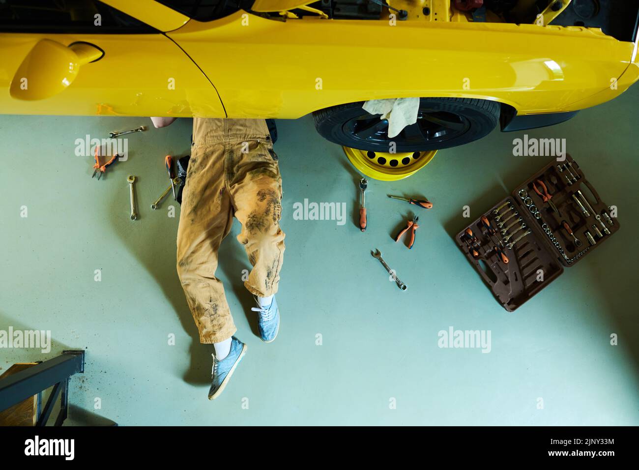 Vista superior del reparador con monos y zapatos de goma debajo del coche amarillo en el suelo del taller o garaje y los detalles de fijación con implementos Foto de stock