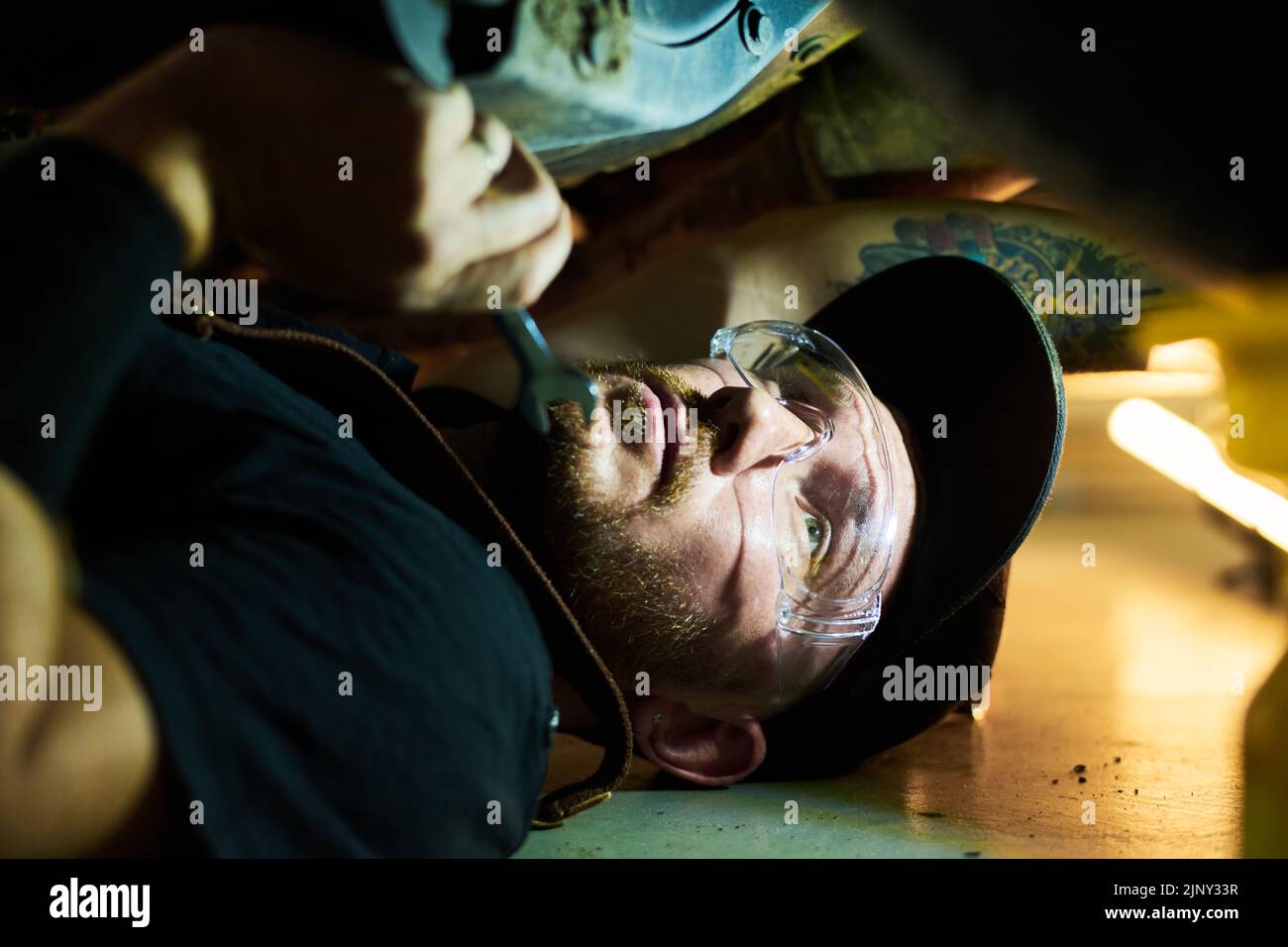 Primer plano de un joven reparador con gafas protectoras y ropa de trabajo debajo del coche mientras estalla su estado y fija los detalles con una llave Foto de stock