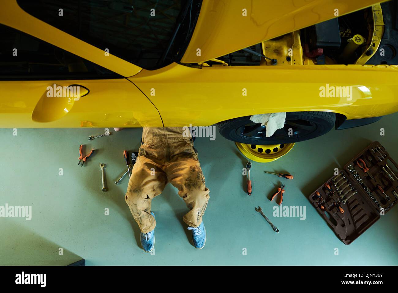 Por encima de la vista de las piernas del mecánico o reparador de los detalles de fijación del coche mientras que acostado en el suelo bajo el automóvil amarillo en el taller Foto de stock