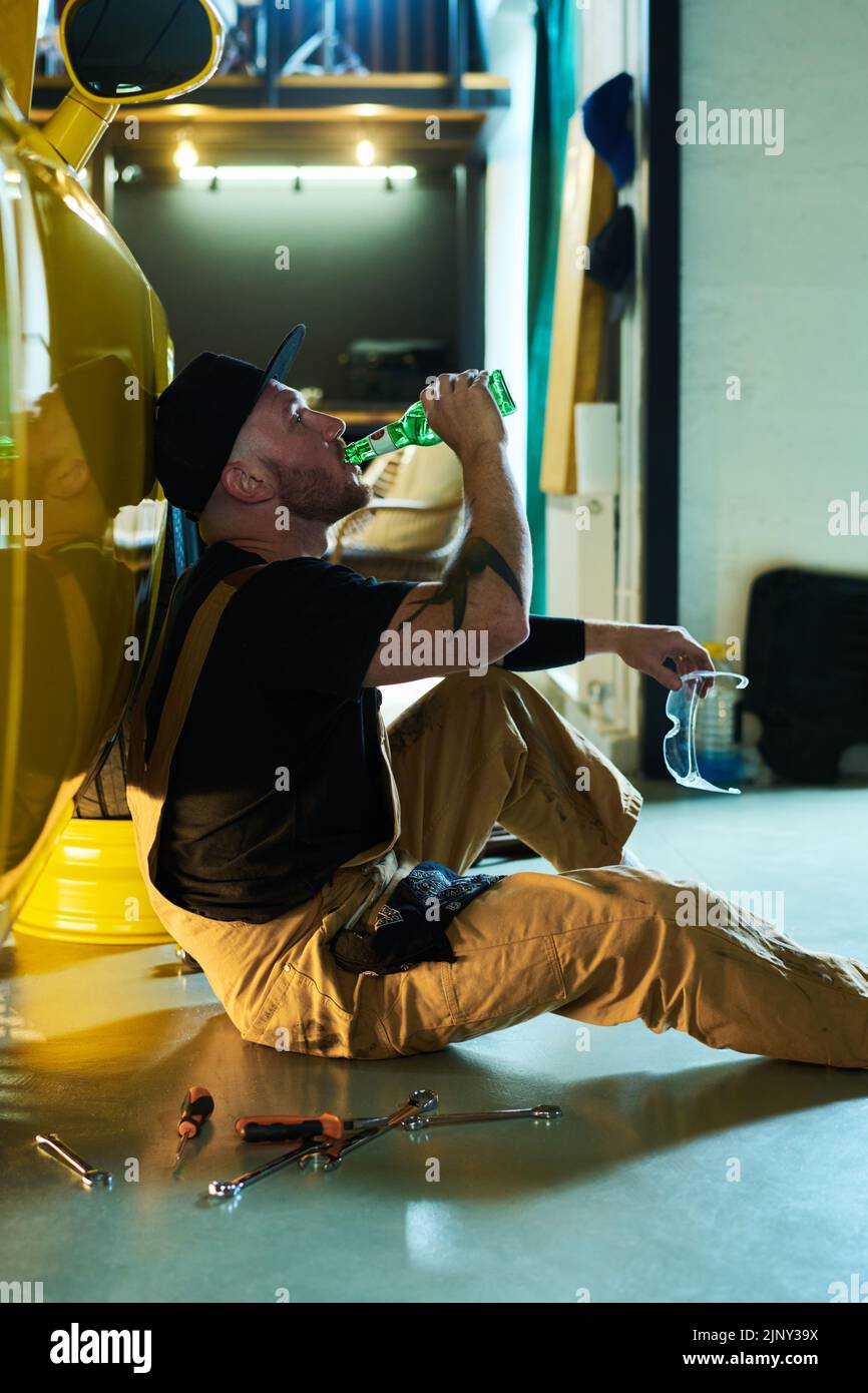 Joven mecánico cansado en ropa de trabajo beber cerveza de botella mientras se sienta en el suelo en coche amarillo después de reparación o revisión técnica Foto de stock