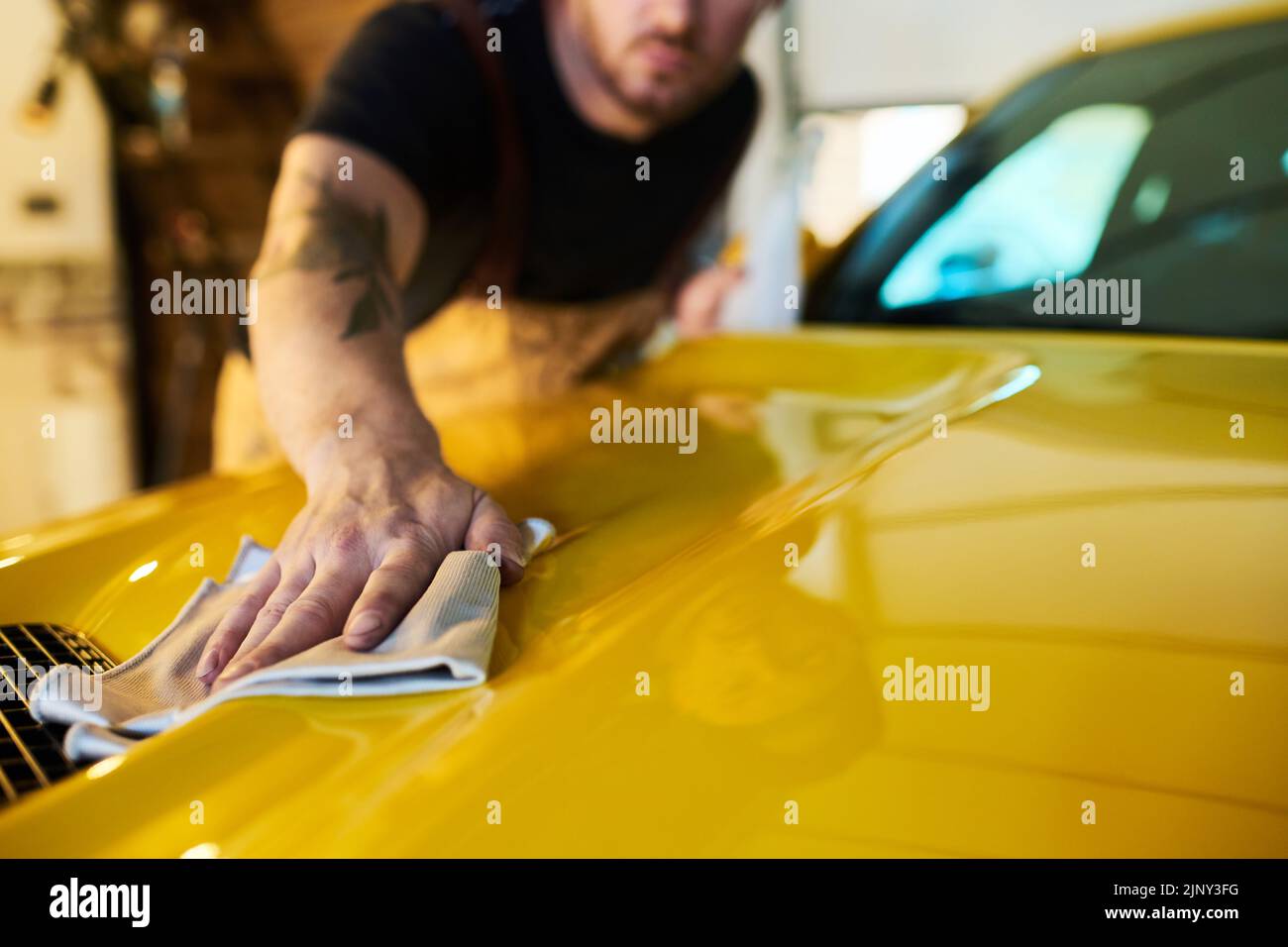 Mano de un técnico joven con un plumero que limpia la superficie de la carrocería amarilla del coche mientras limpia la cubierta del capó después de la reparación Foto de stock