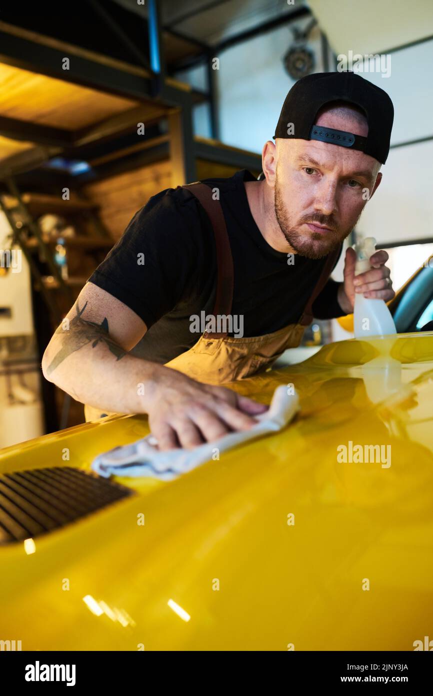Mecánico joven o técnico de mantenimiento, servicio de limpieza de la cubierta del capó con detergente mientras se dobla sobre la parte delantera de la carrocería del coche Foto de stock