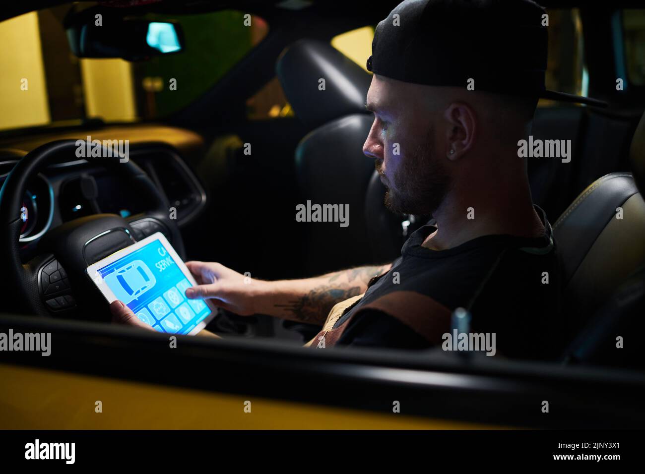 Técnico o mecánico joven con tableta digital mirando los iconos en la pantalla del gadget mientras se sienta delante del volante en el coche Foto de stock