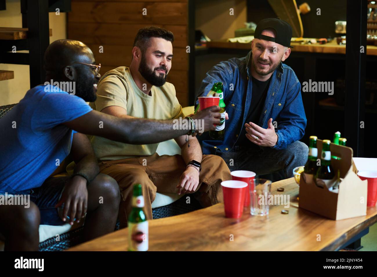 Grupo de jóvenes amigos interculturales clinking con cerveza sobre la mesa con tazas y alcohol mientras disfruta de su reunión en el garaje Foto de stock