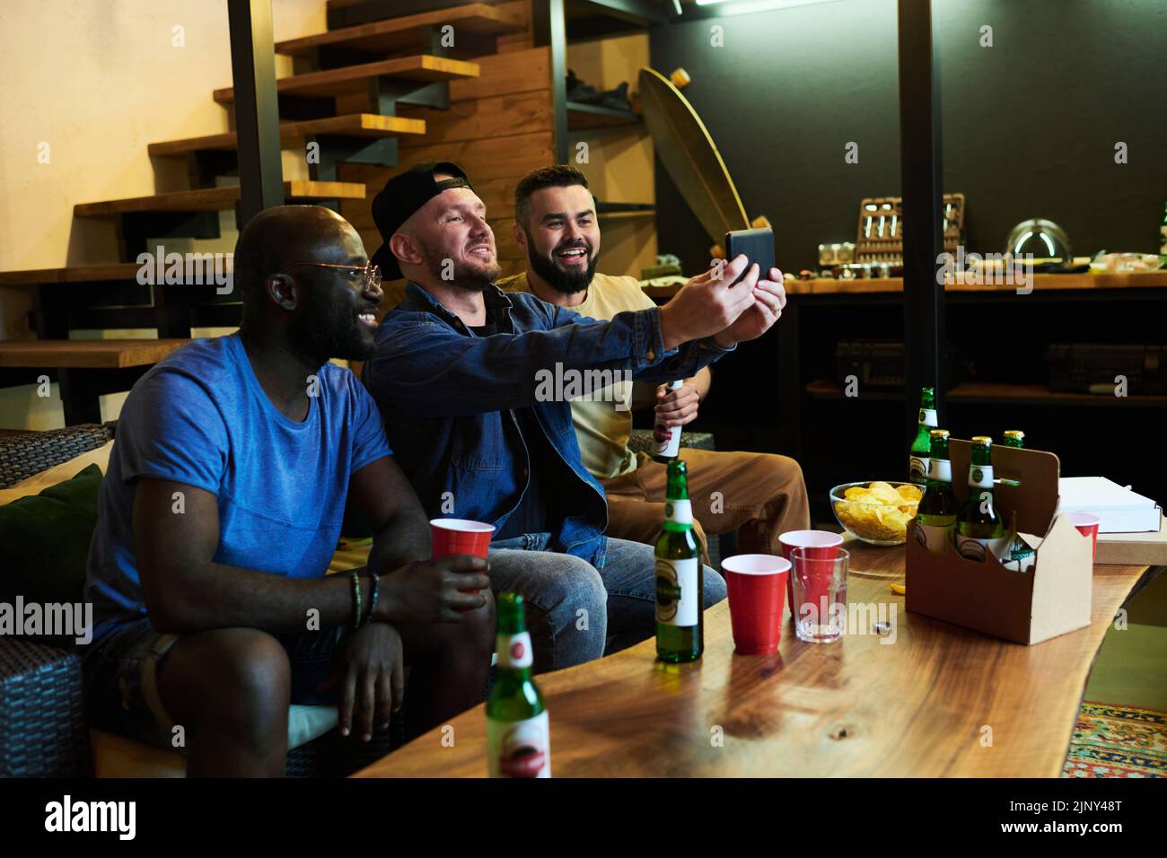 Tres jóvenes amigos interculturales felices haciendo selfie en smartphone mientras se relajan en el sofá frente a la mesa con cerveza y aperitivo Foto de stock
