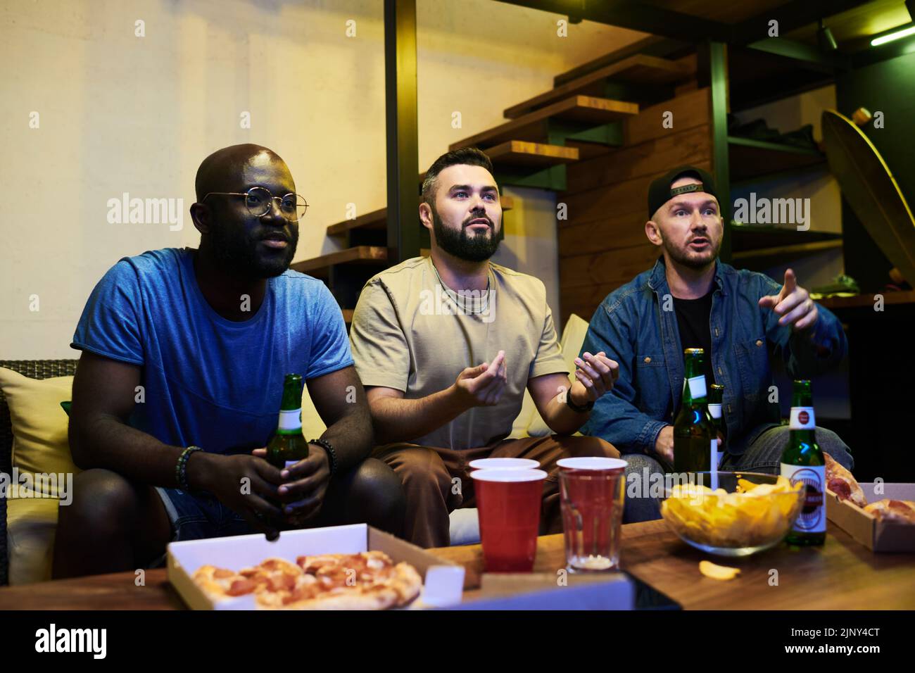 Grupo de jóvenes amigos interraciales viendo la emisión de un partido de fútbol en el garaje sentado frente a una mesa servida y un televisor Foto de stock