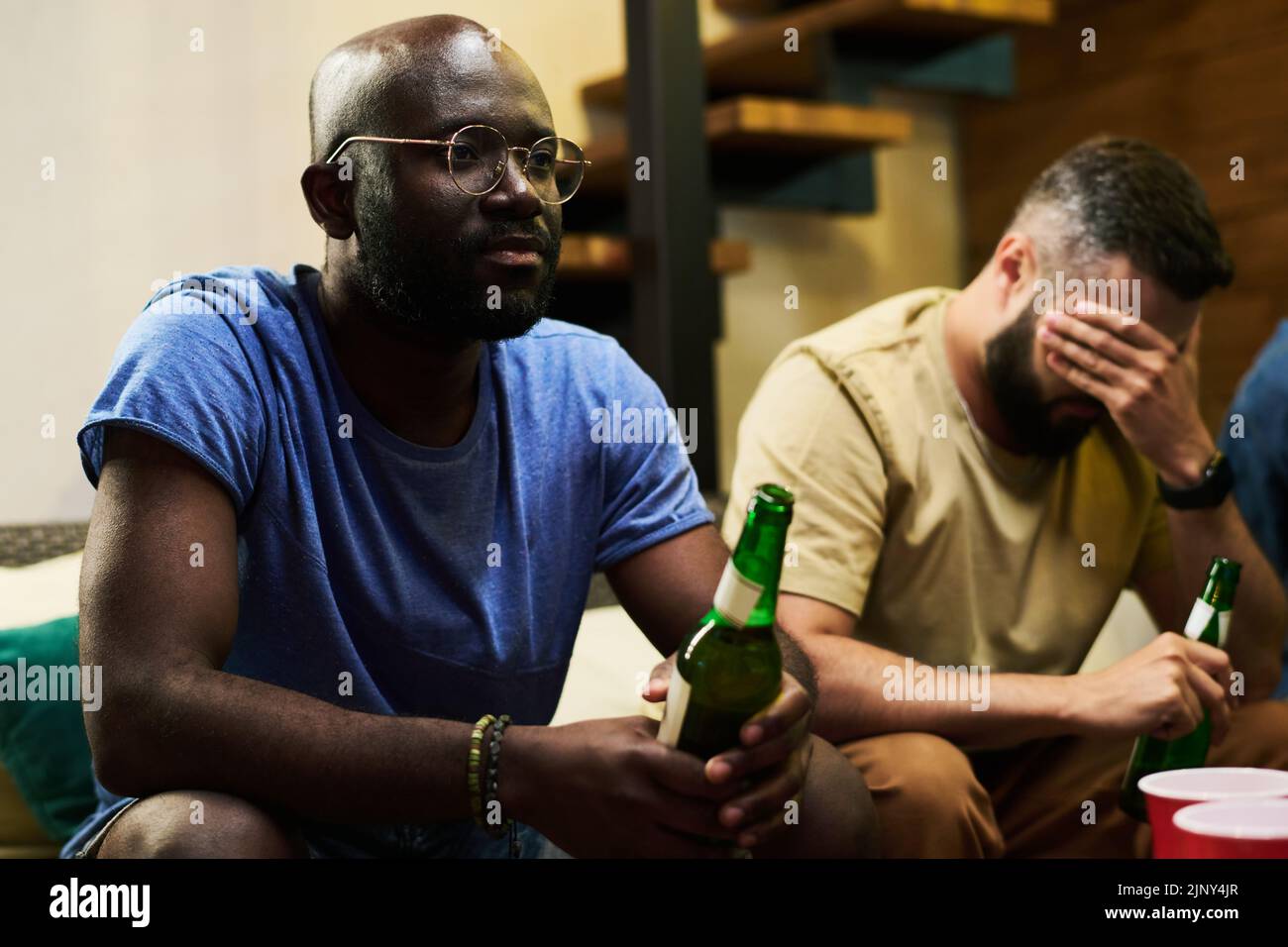 Joven hombre afroamericano disgustado con una botella de cerveza viendo la emisión de televisión de un partido de fútbol con sus amigos en el garaje Foto de stock