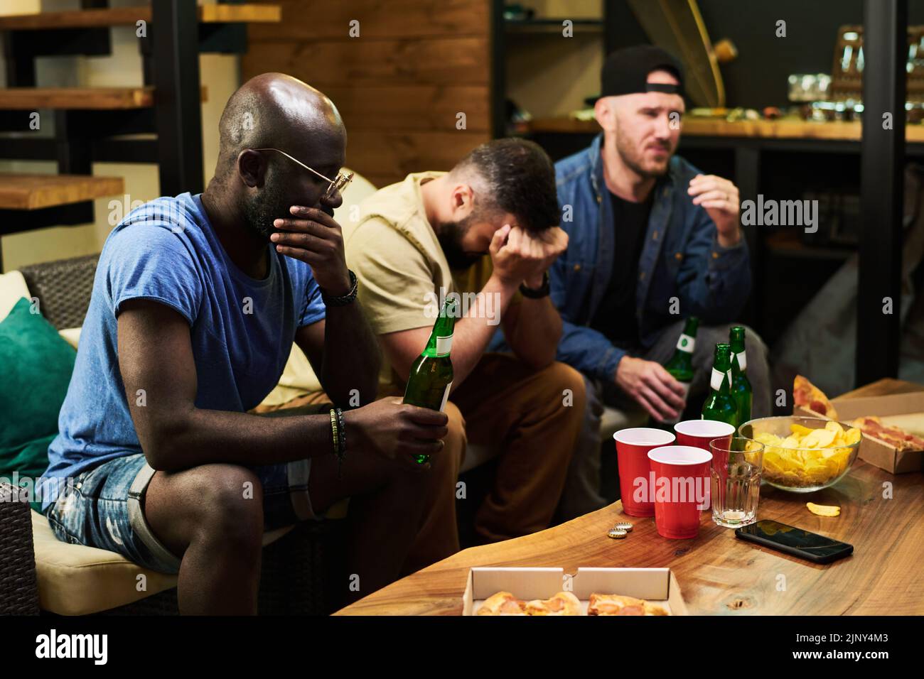 Grupo de jóvenes amigos molestos que expresan tristeza por su equipo de fútbol favorito mientras se sientan delante de la televisión y ven la emisión Foto de stock