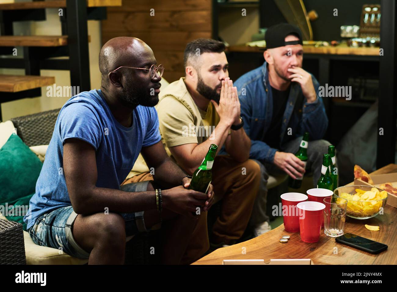 Tres jóvenes amigos interculturales que expresan preocupación mientras ven la emisión del partido de fútbol delante de la mesa con cerveza y merienda Foto de stock