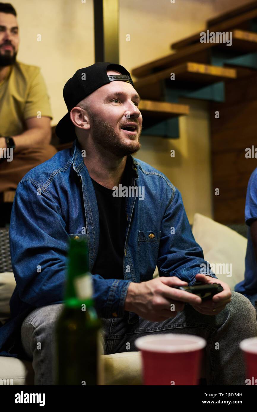 Hombre joven feliz con chaqueta vaquera y gorra presionando los botones del gamepad mientras está sentado en el sofá frente al televisor y jugando a videojuegos Foto de stock