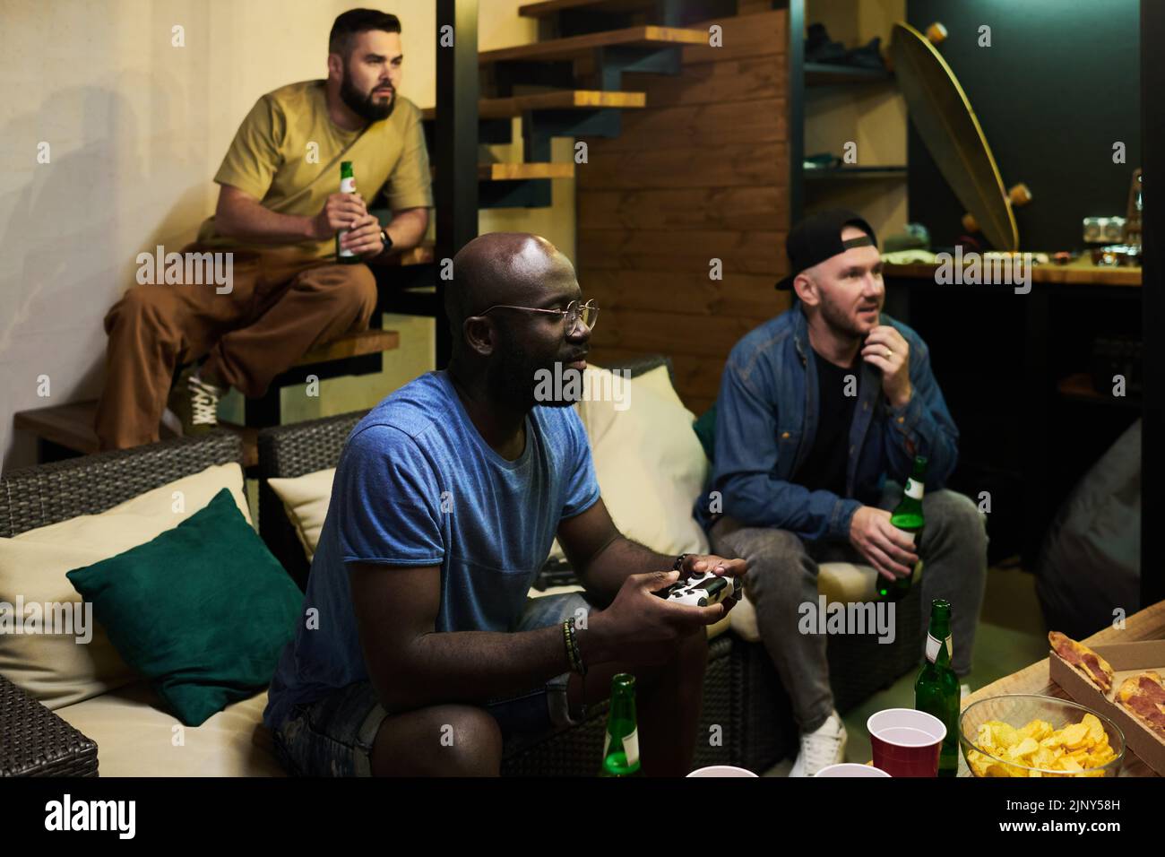 Dos hombres jóvenes con cerveza mirando la pantalla de televisión y viendo un juego de vídeo mientras que el hombre negro con joystick cerca y pulsando botones Foto de stock
