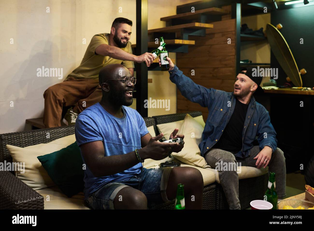 Dos chicos golpeando con botellas de cerveza mientras su amigo afroamericano con joystick sentado en el sofá y jugando a videojuegos Foto de stock