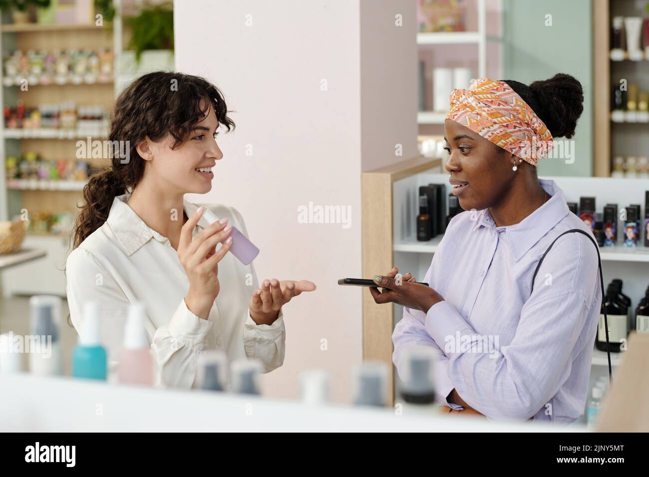 Joven asistente de tienda que recomienda un producto cosmético mujer negra en una pequeña botella de plástico con pulverizador en el supermercado Foto de stock