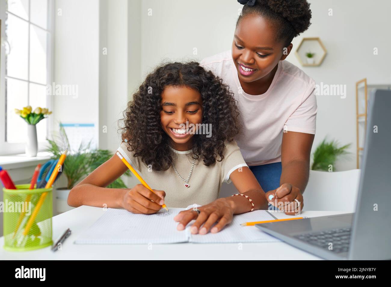 Adolescente afroamericana alegre haciendo la tarea escolar en casa con la ayuda de mamá. Foto de stock