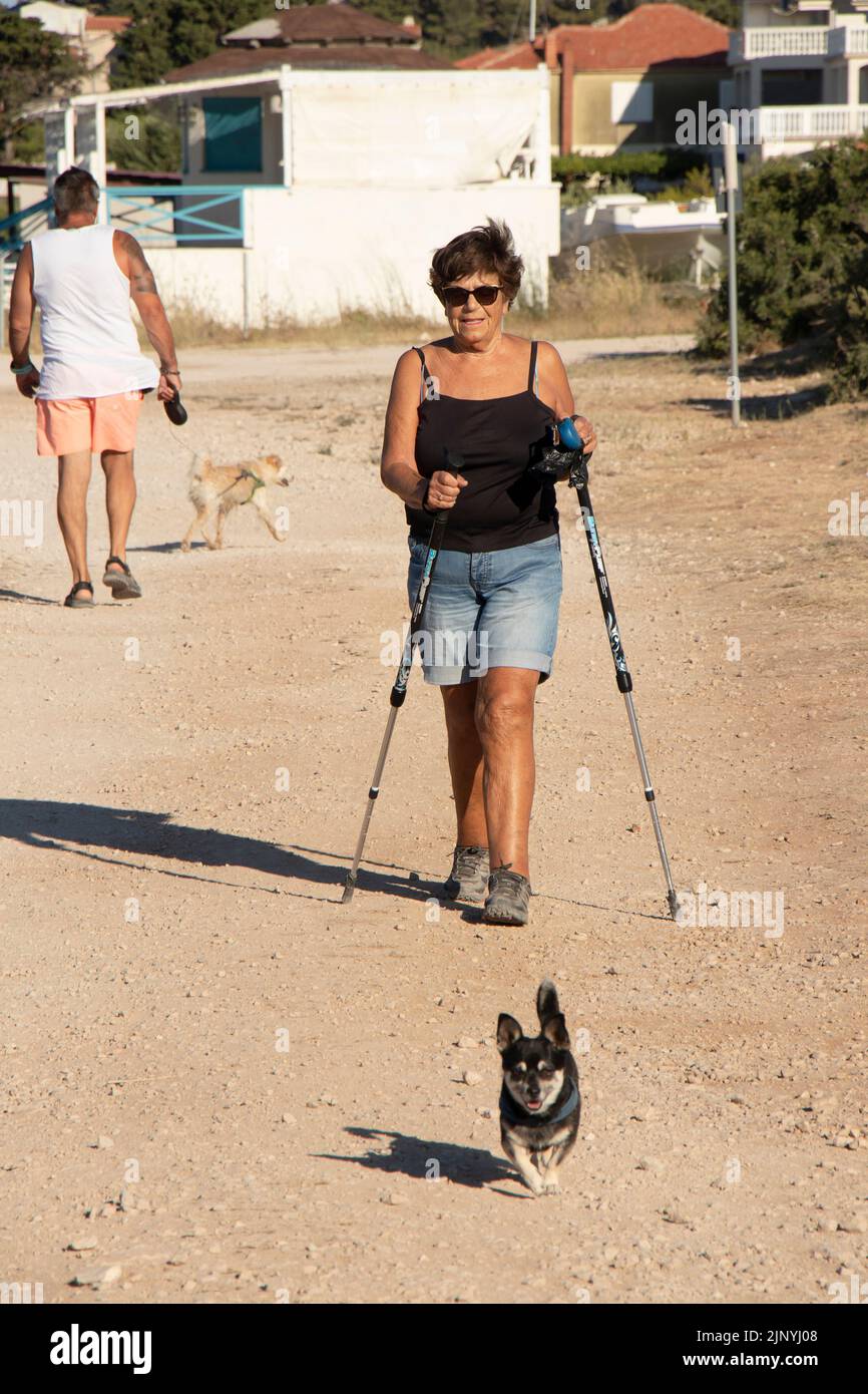 Vodice, Croacia - 13 de julio de 2022: Mujer anciana caminando con palos de caminar con su perro en una pasarela de tierra de la costa Foto de stock