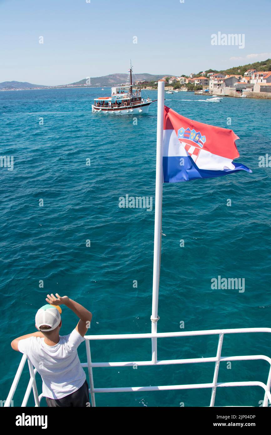 Isla de Prvic, Croacia - 22 de julio de 2022: Un niño ondeando desde el barco con bandera croata para hacer una excursión diaria en el casco antiguo de Prvic, trasera vie Foto de stock