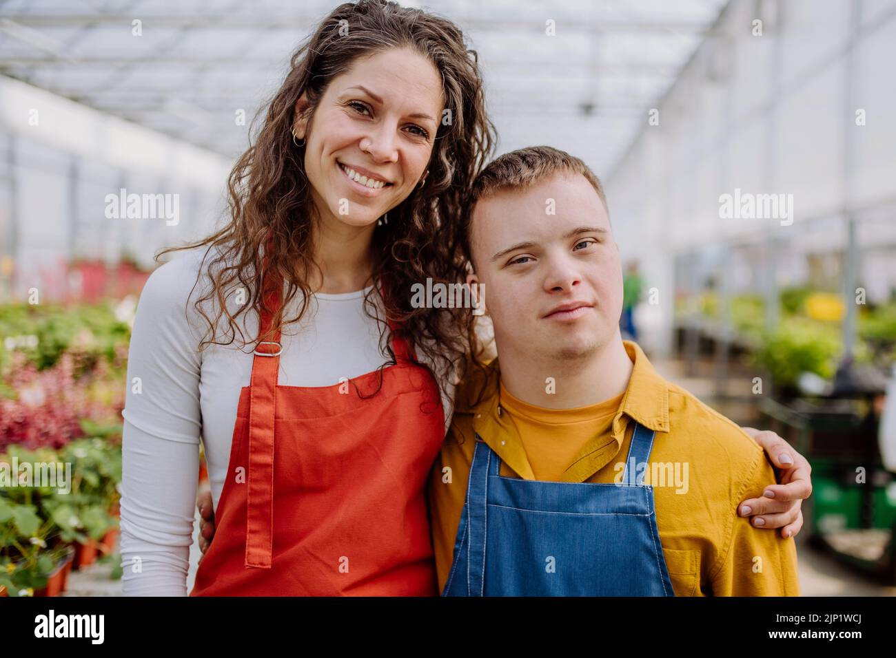 Florista mujer experimentada posando con colega joven con síndrome de Down en el centro de jardín. Foto de stock