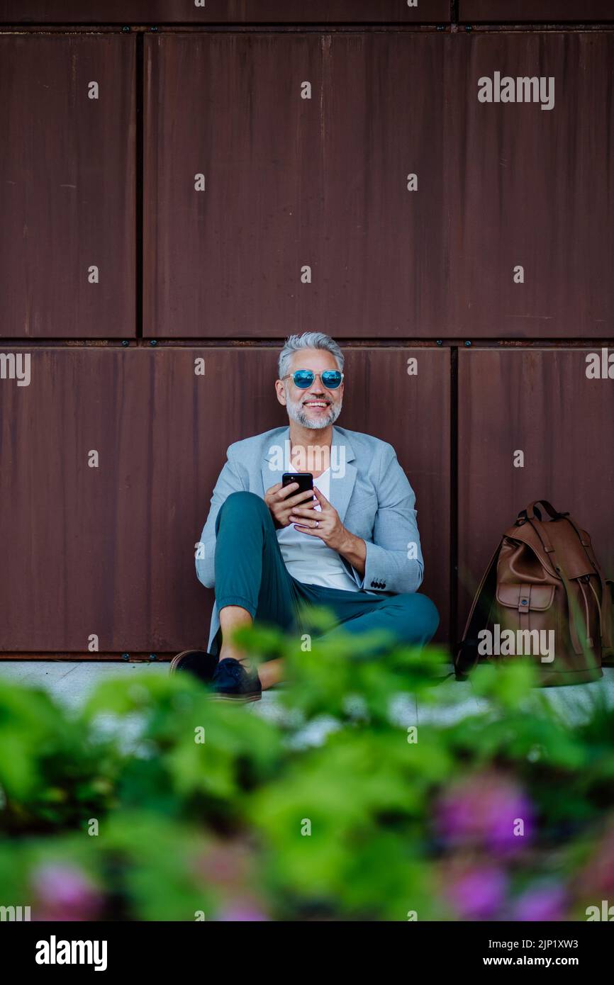 Hombre de negocios maduro sentado con un smartphone en la mano y relajándose en el parque de flores de la ciudad durante un descanso en el trabajo. Foto de stock