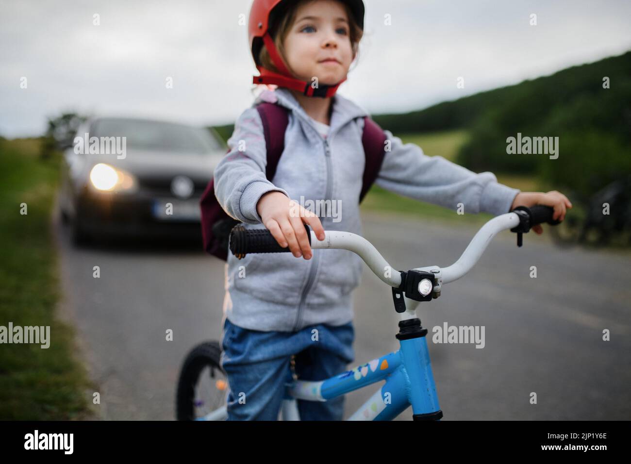 Retrato de una niña emocionada montando en bicicleta por carretera con el coche detrás de ella, concepto de educación en seguridad vial. Foto de stock