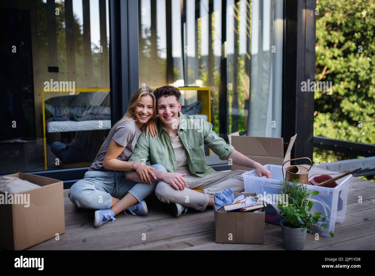 Alegre joven pareja mudarse en su nueva y diminuta casa en el bosque sentado en el suelo en la terraza. Concepción del movimiento y la vida sostenible. Foto de stock