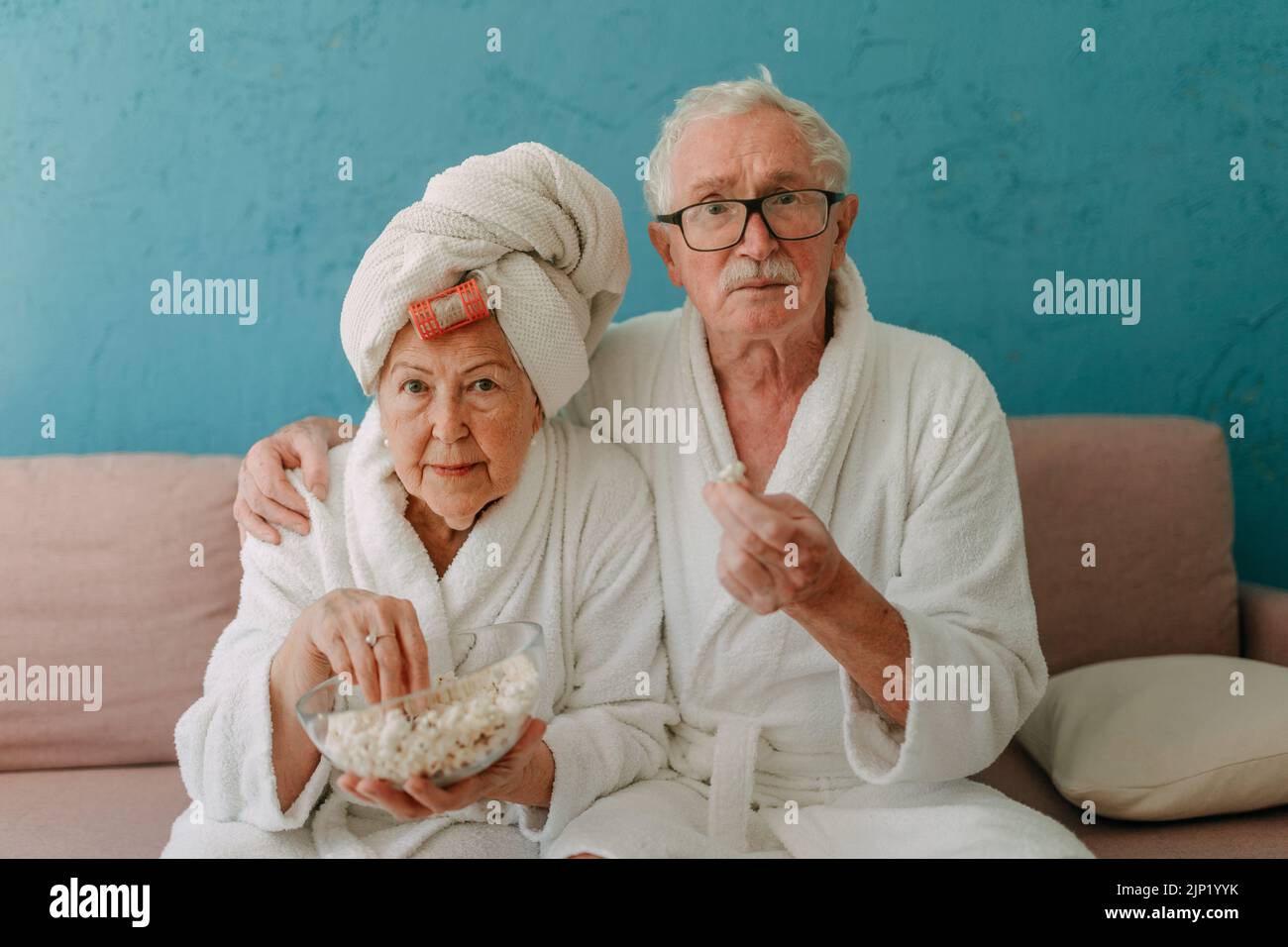 Una feliz pareja mayor sentada en el sofá en los albornoces y viendo la televisión con palomitas de maíz. Foto de stock