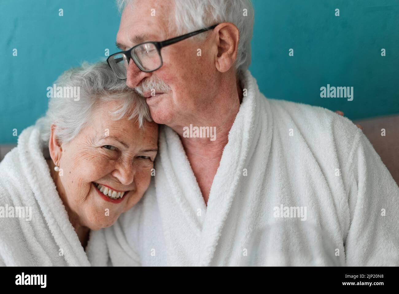 Retrato de una feliz pareja mayor sentada en un albornoz en el sofá, pasándolo bien en casa. Foto de stock
