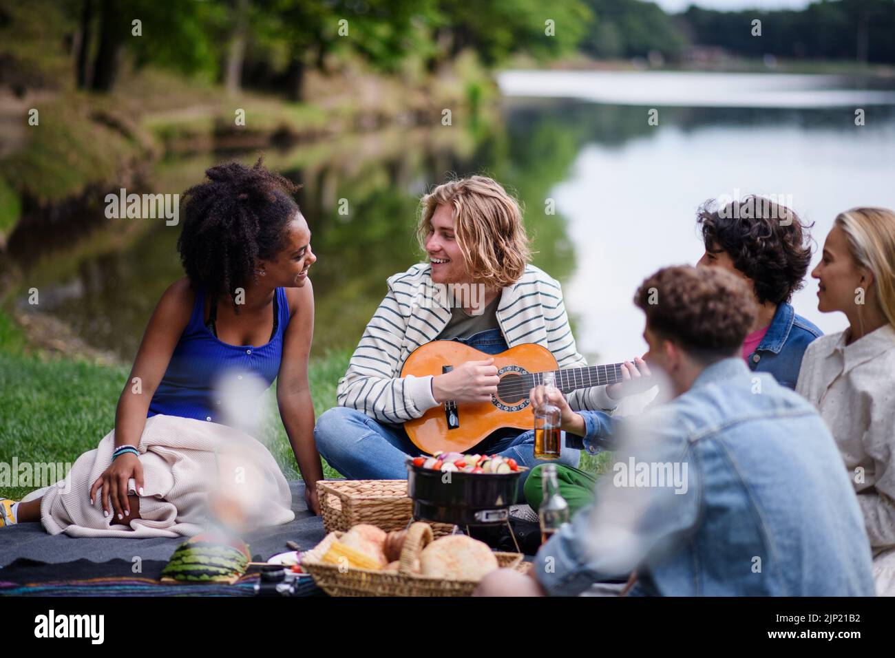 Grupo de amigos divirtiéndose en un picnic cerca de un lago, sentados en la manta comiendo y tocando la guitarra. Foto de stock