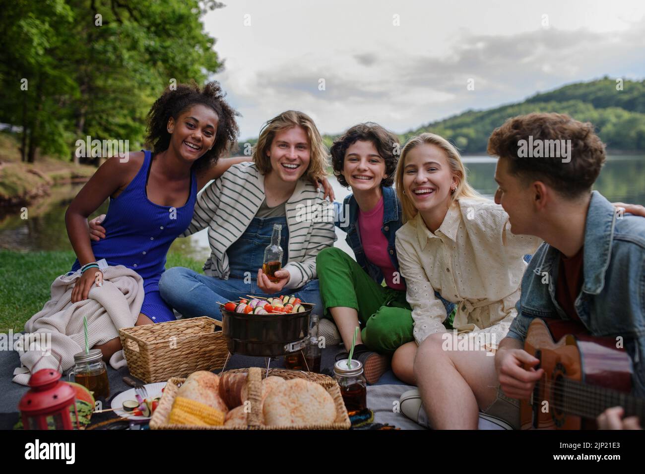 Grupo de jóvenes amigos multirraciales acampando cerca del lago y haciendo barbacoas juntos, mirando la cámara. Foto de stock
