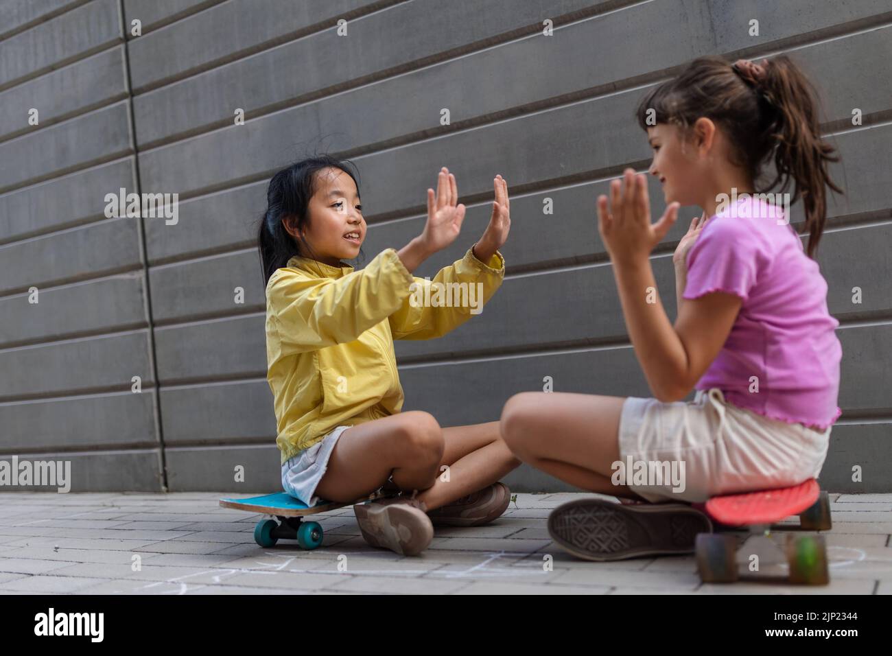 Chica asiática con su amiga sentada en patinetas en la calle de la ciudad, hablando y jugando, concepto de estilo de vida activo de los niños. Foto de stock