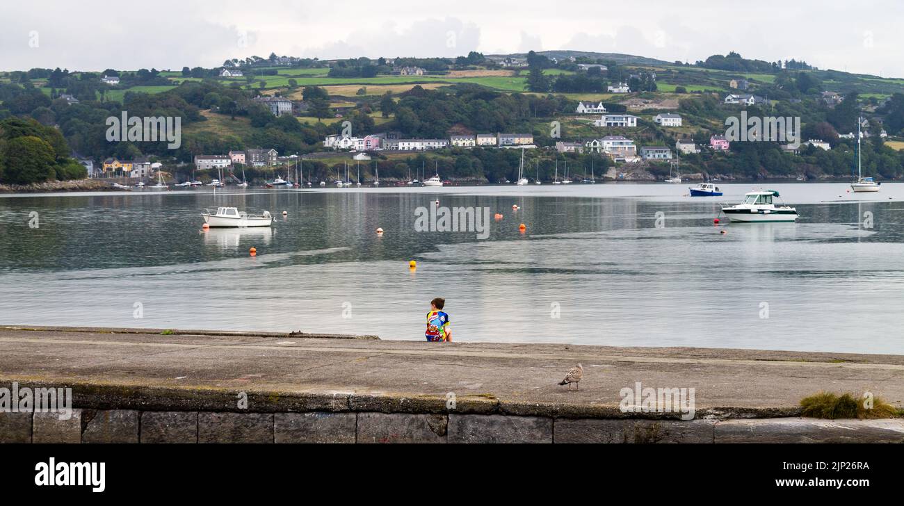 Niño pequeño sentado solo en soledad en la pared del puerto mirando al mar. Foto de stock