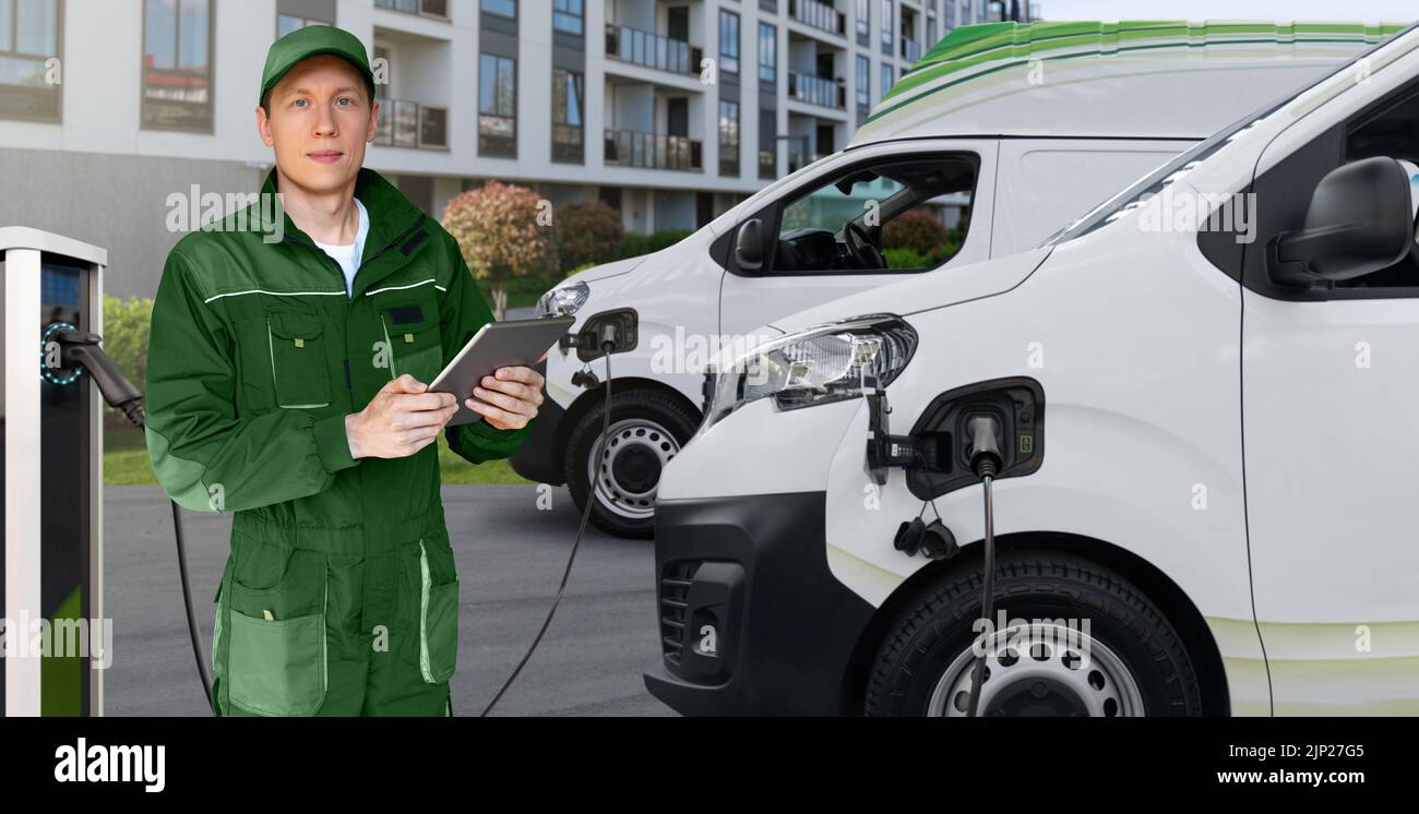 Un hombre con una tableta digital se encuentra junto a los furgones de reparto eléctrico en las estaciones de carga de vehículos eléctricos Foto de stock