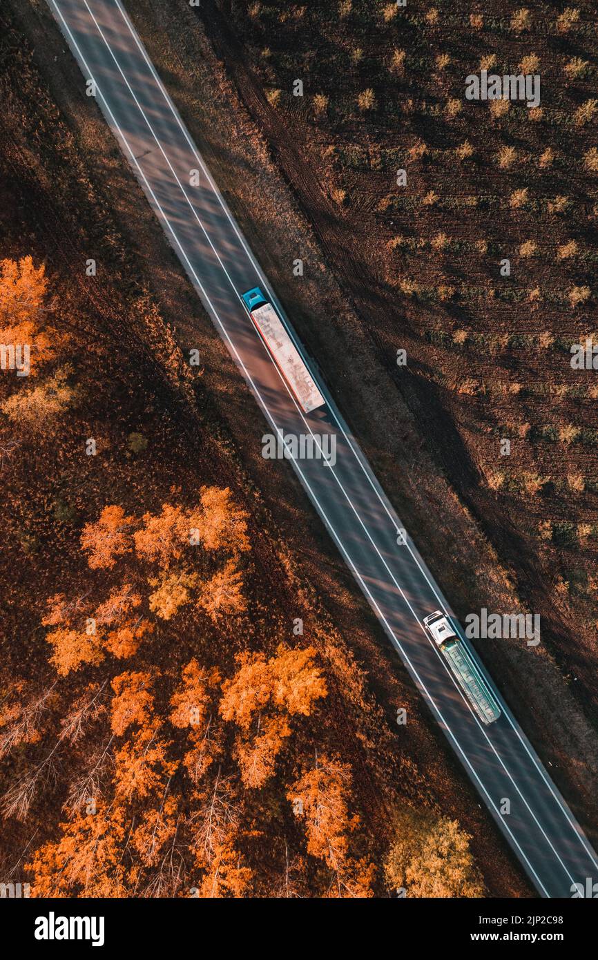 Foto aérea de dos camiones en la carretera a través de bosque caducifolio en la tarde de otoño, vista de arriba hacia abajo de drone pov de los vehículos en la carretera Foto de stock